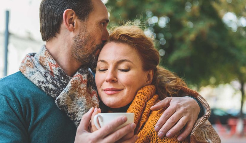 man kissing woman forehead outside with mug